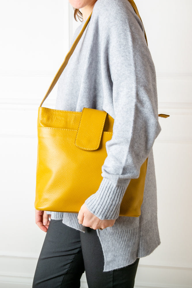 A model wearing the Pimlico Handbag in mustard yellow colour, shown from the front and including the adjustable leather strap, the fold over presstud fastening and the outside pocket. Made from Italian leather.
