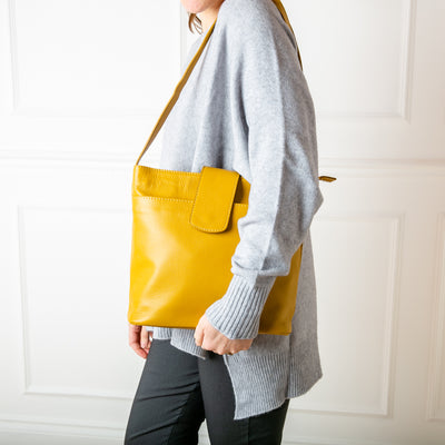 A model wearing the Pimlico Handbag in mustard yellow colour, shown from the front and including the adjustable leather strap, the fold over presstud fastening and the outside pocket. Made from Italian leather.
